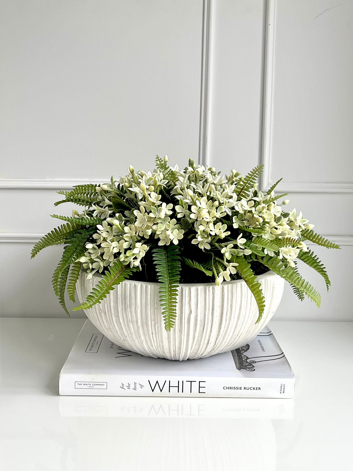 Artificial white jasmine and lush green ferns surrounded by preserved moss in a white ribbed ceramic bowl planter displayed on a book.