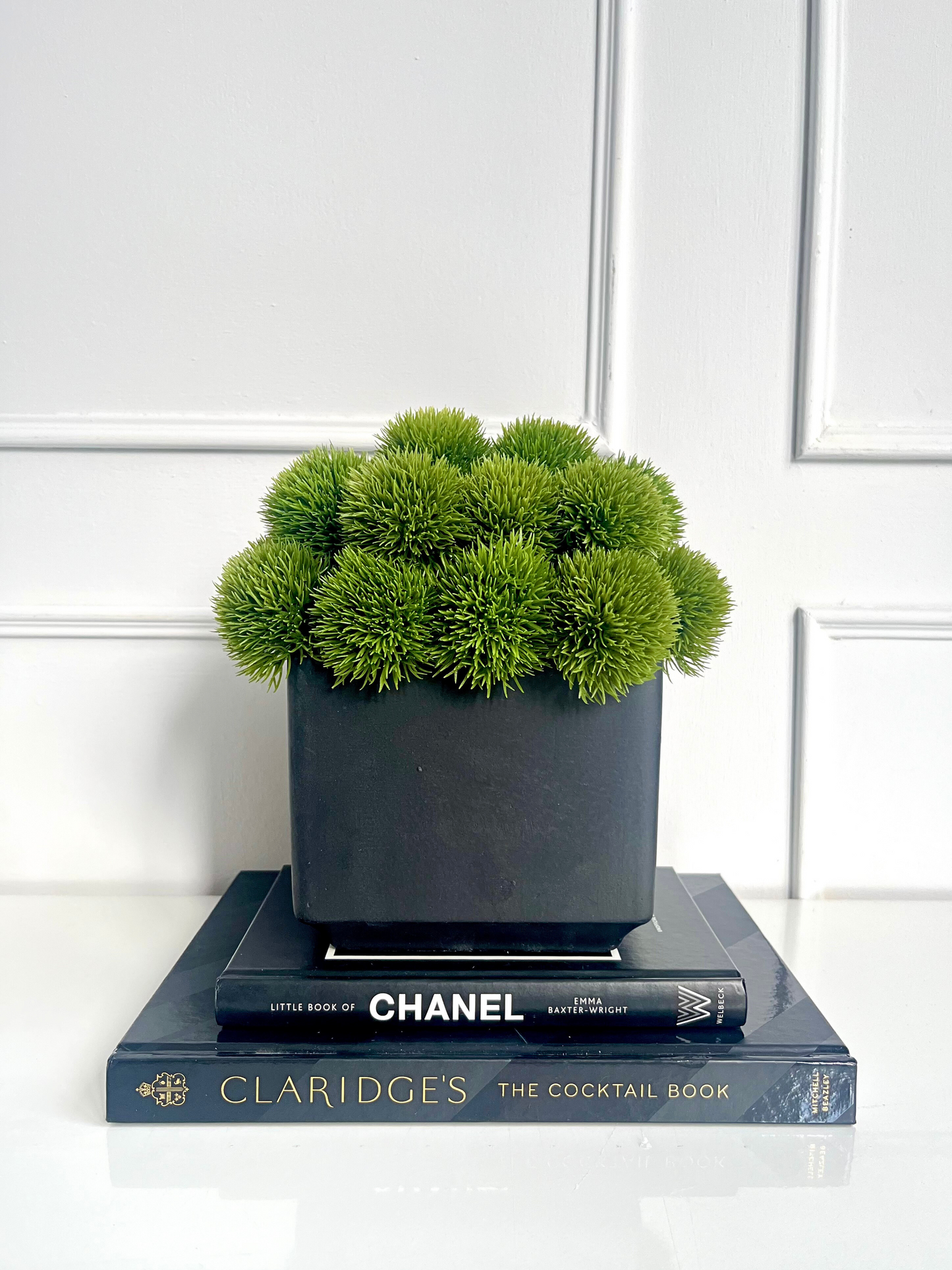 Artificial lush green dianthus set in a matt black cube planter displayed on a stack of books.