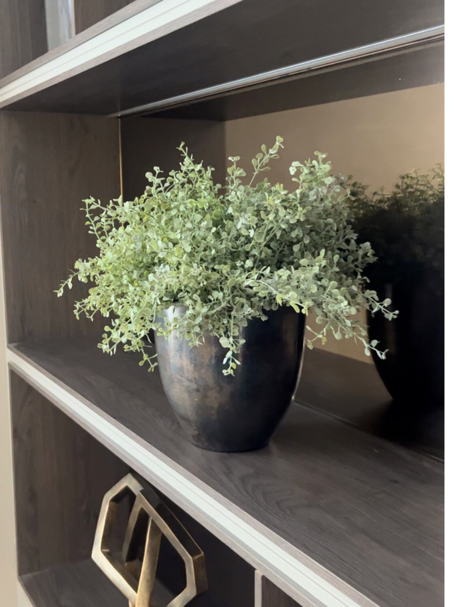 Soft green artificial button leaf plant set in an aluminium bronze coloured pot displayed on a media unit shelf.