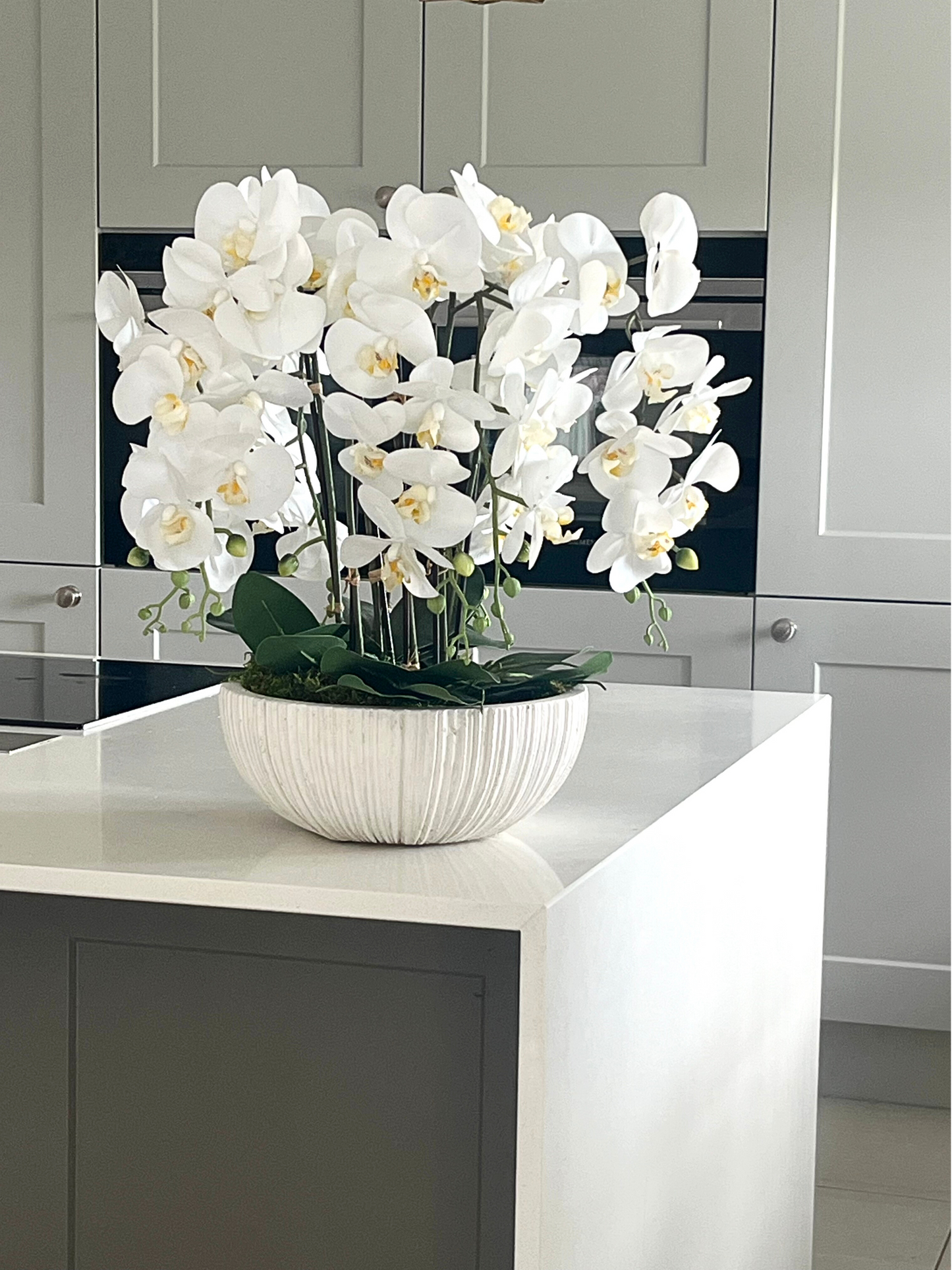 Artificial white orchids set in a white ribbed bowl planter in preserved moss displayed on a kitchen island with grey cupboards and a white waterfall worktop.