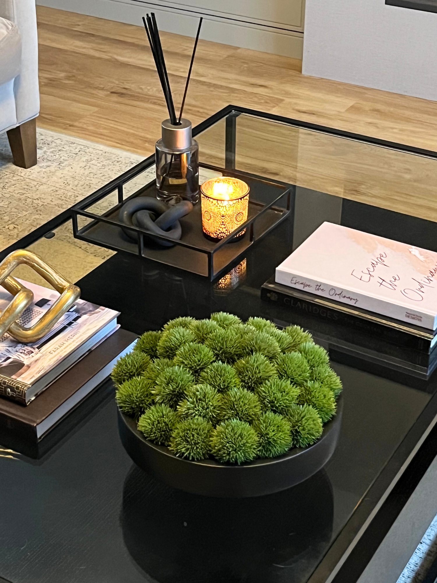 Artificial vibrant green dianthus set in a black wooden bowl displayed on a coffee table with books, an ornament, tray, diffuser and candle.