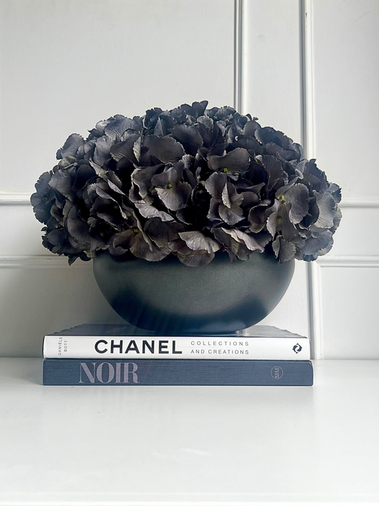 Dark grey artificial hydrangea set in a matt black ceramic bowl planter displayed on a stack of books.