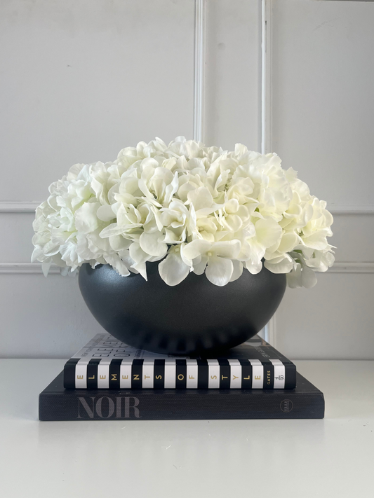 white artificial hydrangea arranged in a black ceramic bowl planter displayed on a stack of books.