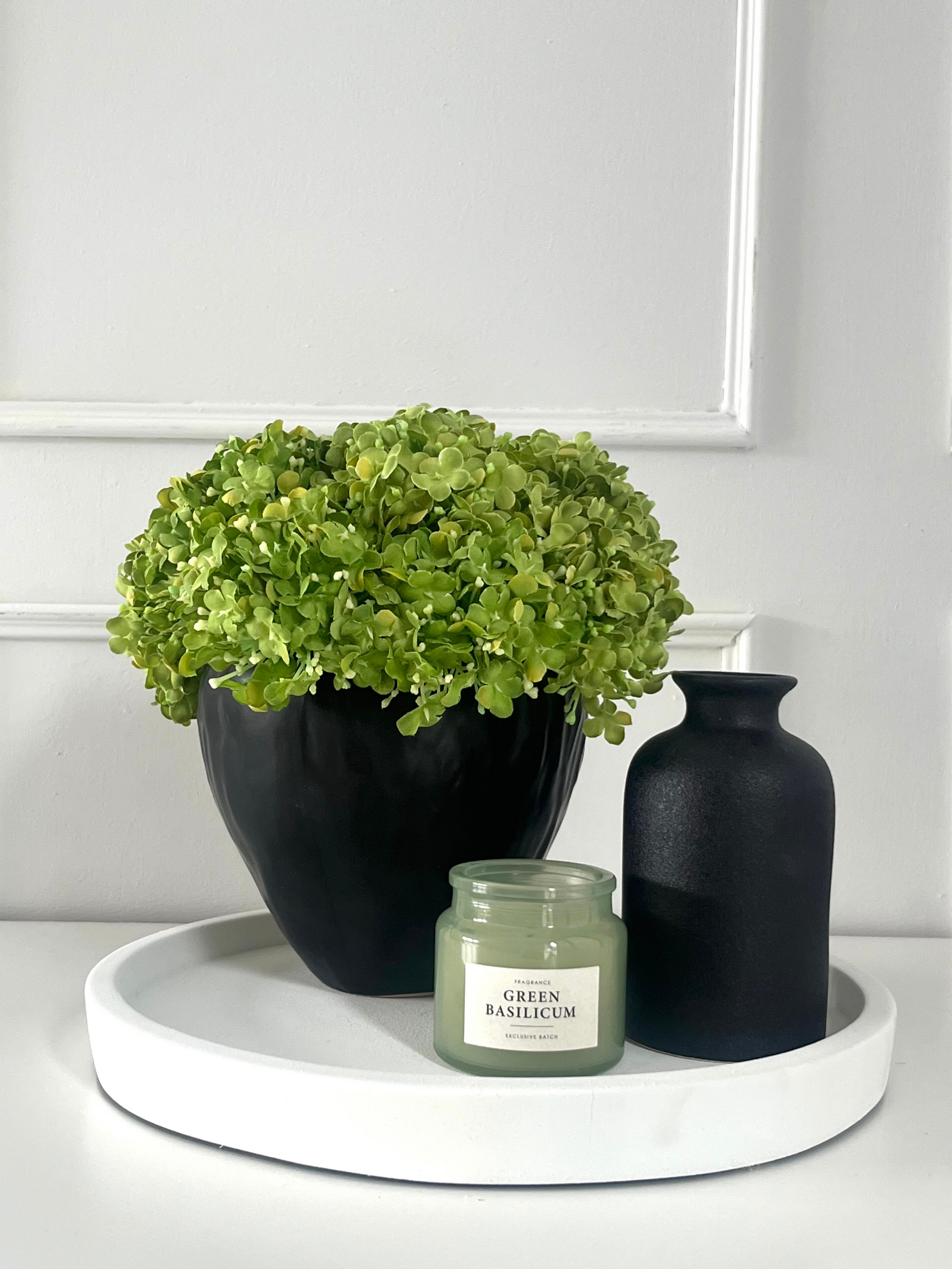 Vibrant green artificial snowball viburnum arranged in a black stoneware pot displayed on a cream tray with a candle and black vase.