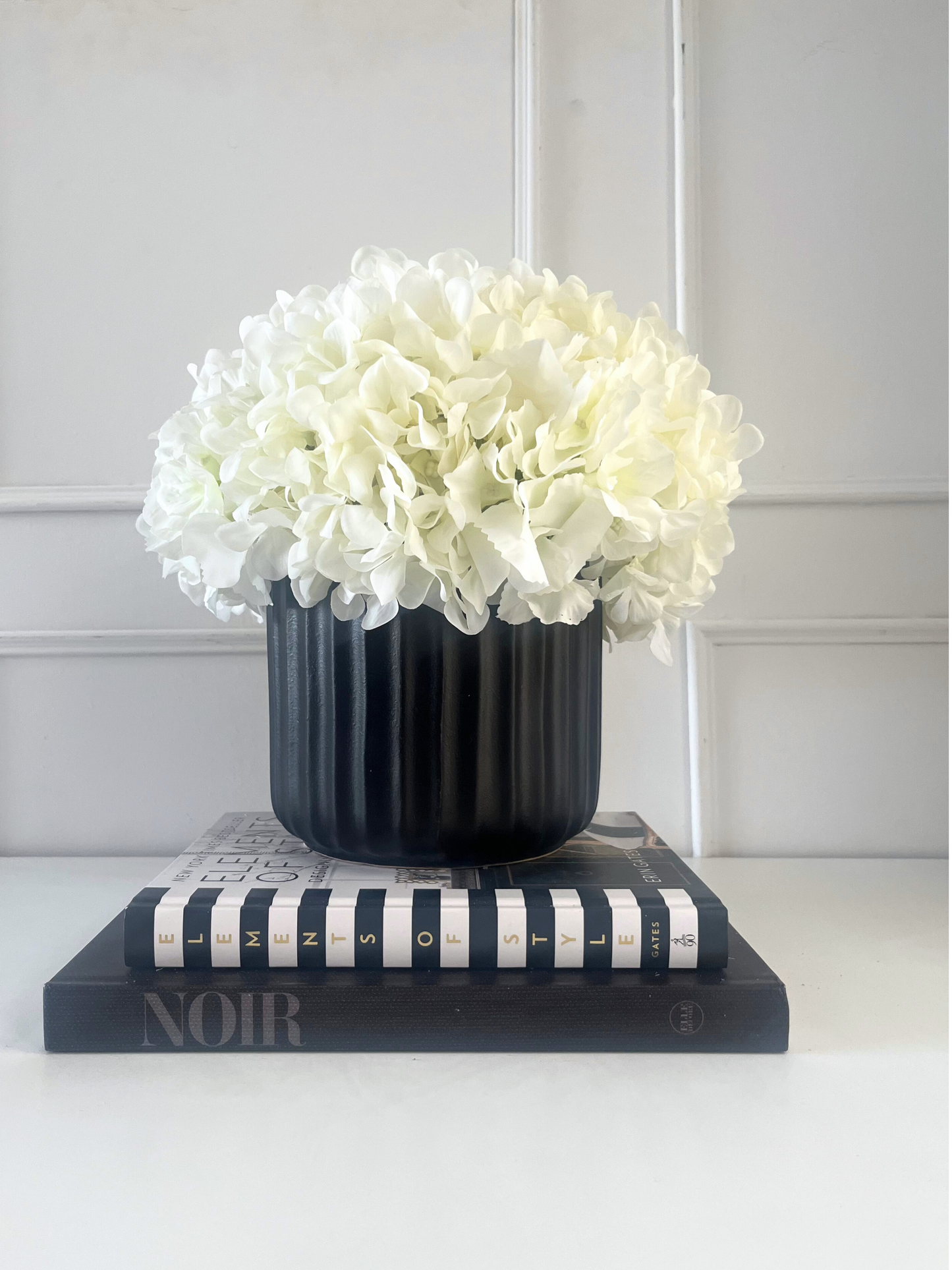 Artificial white hydrangea set in a black porcelain ribbed pot displayed on a stack of books.