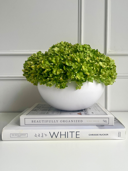 vibrant green artifiical snowball viburnum arranged in a crisp white ceramic bowl planter displayed on a stack of books.