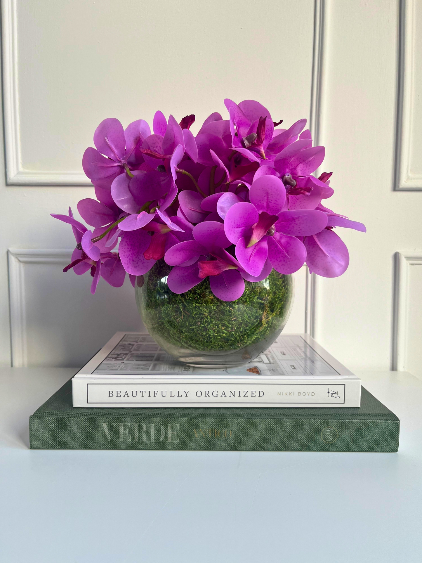 Artificial pink orchids set in a preserved moss lined glass ball vase displayed on a stack of books.